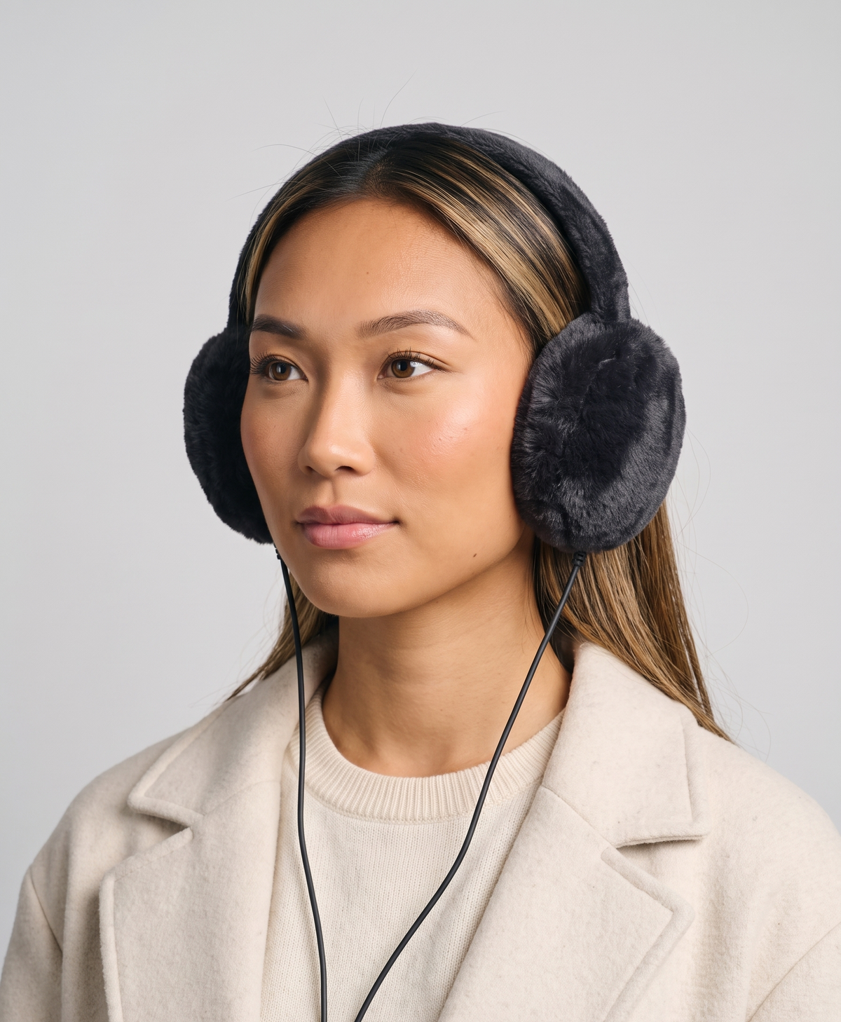 Gray plush heated earmuffs for women on a gold wire stand, with the connector cable and a power bank in front