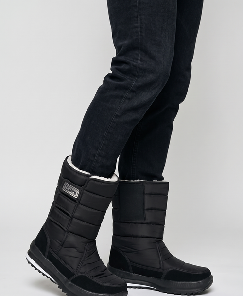 Man wearing black quilted heated snow boots with jeans, standing in deep snow