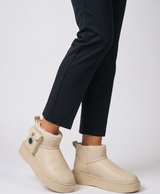 Woman wearing beige fleece-lined heated snow boots with a side pocket and power button, seated on a cream sofa