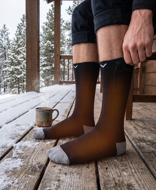 Black heated socks with 360° heat graphic over a snowy mountain slope with a snowboarder