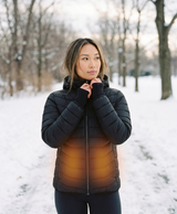 Women's black heated puffer jacket with hood, front zip and chest power button, shown with heat zones lit